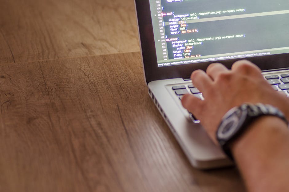 A person working at a wooden desk with a laptop displaying lines of code on its screen, positioned centrally in the image. To the left of the laptop is a black camera with an attached zoom lens, stand