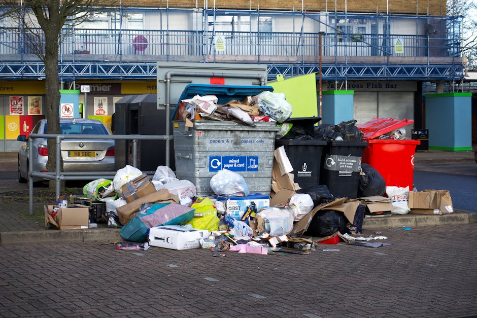A collection of overflowing waste bins and scattered rubbish on a paved urban street in front of a building with a blue scaffolding structure. The scene includes a large gray mixed paper and cardboard