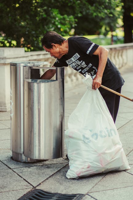A collection of overflowing waste bins and scattered rubbish on a paved urban street in front of a building with a blue scaffolding structure. The scene includes a large gray mixed paper and cardboard