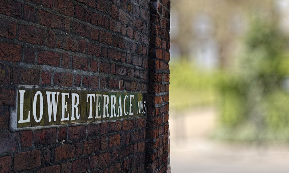 Close-up view of a metal street sign mounted on a textured, red brick wall. The sign reads 'Lower Terrace NW3' with white capital letters on a background of green and white tiles, displaying slight we